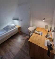 Ein einfaches Zimmer mit einem Einzelbett und einem kleinen Schreibtisch. Die Wände sind hell, und es gibt eine Schreibtischlampe sowie einen Stuhl.