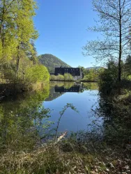 Ein ruhiger Teich umgeben von Bäumen und sanften Hügeln. Im Hintergrund ist ein Gebäude sichtbar, das sich im Wasser spiegelt.