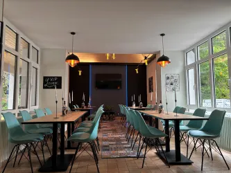 Ein stilvoller Raum mit langen Tischen und blaugrünen Stühlen. Große Fenster lassen viel Licht herein, und die Wand hat einen modernen Fernseher.