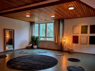 Ein heller Raum mit Holzdecke und großen Fenstern. Auf dem Boden liegen runde Teppiche und es steht eine Pflanze in der Ecke.
