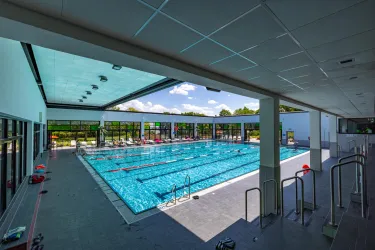 Ein helles Hallenbad mit einem großen Schwimmbecken. Die Fenster bieten einen Blick ins Freie und auf blauen Himmel.