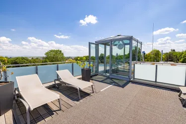 Eine moderne Dachterrasse mit Liegen und einem Glasaufzug. Der Himmel ist klar und blau, umgeben von viel Grün.