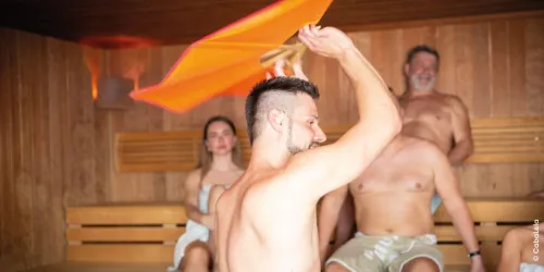 Eine Sauna mit mehreren Personen, die sich entspannen. Ein Mann schwingt ein Handtuch, um Luftzirkulation zu erzeugen.