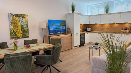 Ein modernes Wohnzimmer mit einer gemütlichen Couch und einem Esstisch. Die offene Küche ist hell und gut ausgestattet, mit einem großen Fernseher in der Ecke.
