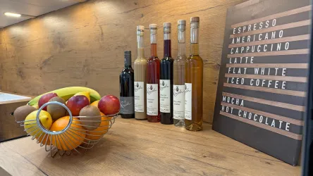 Eine moderne Theke mit einer Auswahl an Getränken in Flaschen steht neben einer dekorativen Obstschale. Auf einem Holzschild sind verschiedene Kaffeespezialitäten aufgelistet.