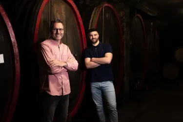 Zwei Männer stehen in einem Weinkeller neben großen Holzfässern. Die Atmosphäre ist gemütlich und einladend.