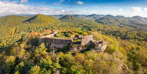 Eine eindrucksvolle Burgruine liegt hoch oben auf einem Hügel. Die Umgebung ist von grünen Wäldern und sanften Hügeln geprägt.