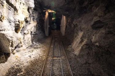 Ein dunkler Stollen mit einer Schienenführung. Der Weg führt zu einer beleuchteten Öffnung im Hintergrund.