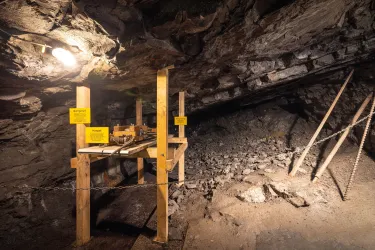 Ein unterirdischer Raum mit Holzgestellen und Werkzeugen. Die Wände sind aus Felsen und die Beleuchtung ist schwach.