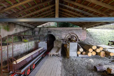 Eine alte Werkstatt mit Holzstücken und Maschinen. Die Umgebung zeigt eine rustikale Atmosphäre unter einem schützenden Dach.