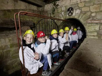 Eine Gruppe von Kindern sitzt in einem Tunnel und trägt Schutzhelme. Sie zeigen freundlich mit der Hand und tragen weiße Laborkittel.