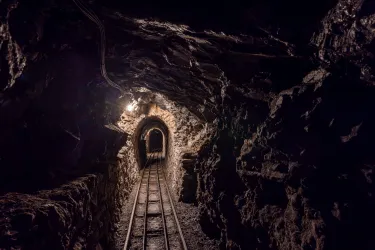 Ein dunkler Stollen mit Gleisen, die durch einen Tunnel führen. An der Decke hängt ein Licht, das den Weg erhellt.