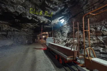Ein unterirdischer Tunnel mit einer Schiene und einer Lorenbahn. Über dem Tunnel hängt ein Schild mit der Aufschrift "Glück Auf".