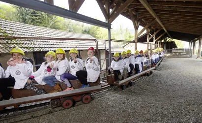 Eine Gruppe von Kindern sitzt in einer kleinen Bahn unter einem Holzdach. Alle tragen gelbe Helme und Warten auf den Beginn der Fahrt.