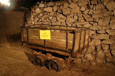 Eine alte Förderwagen steht vor einer Steinmauer in einem Bergwerk. Der Wagen ist aus Holz und hat einen gelben Hinweiszettel.