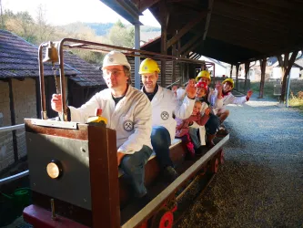 Eine Gruppe von fünf Personen sitzt auf einer kleinen Lokomotive in einer Werkstatt. Alle tragen Sicherheitshelme und lachen fröhlich in die Kamera.