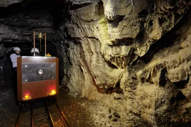 Ein Bergwerk mit einem kleinen Waggon auf Schienen. Im Hintergrund sind graue Felsen mit Stalaktiten sichtbar.