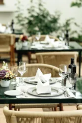 Ein eleganter Tisch in einem Restaurant, dekoriert mit feinem Geschirr und Gläsern. Im Hintergrund sind weitere Tische mit Pflanzenarrangements zu sehen.