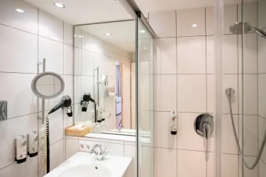 Ein modernes Badezimmer mit einer Dusche und einem Waschbecken. Die Wände sind hell gefliest und es gibt Spiegel und Pflegeprodukte.