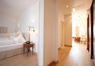 Ein moderner Hotelzimmerbereich mit einem Bett und einem kleinen Tisch. Der Flur führt zu einem hellen Sitzbereich mit Fenster.