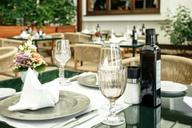 Ein eleganter Tisch in einem Restaurant im Freien mit Glasgeschirr und Pflanzen im Hintergrund. Auf dem Tisch stehen Weingläser, eine Flasche Öl und eine kleine Blumendekoration.