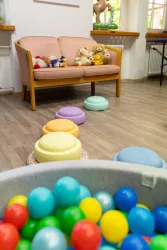 Ein heller Raum mit einem rosa Sofa und einem Spielbereich. Bunte Bälle liegen auf dem Boden und pastellfarbene Kissen sind verteilt.