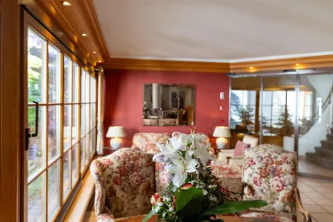 Ein gemütliches Wohnzimmer mit floralem Möbeldesign und großen Fenstern. Im Vordergrund steht ein Tisch mit einer Blumendekoration.