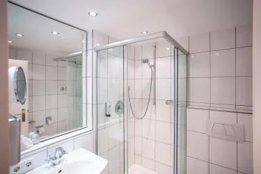 Ein modernes Badezimmer mit einer gläsernen Dusche und weißen Fliesen. Ein Waschbecken und ein Spiegel sind ebenfalls sichtbar.