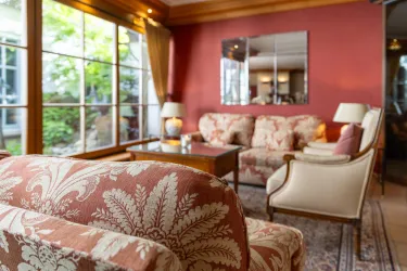 Ein gemütliches Wohnzimmer mit einer großen Fensterfront und Blick ins Grüne. Die Möbel haben ein elegantes Design mit einer warmen Farbpalette.