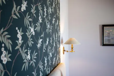 Ein elegantes Zimmer mit einer floral gemusterten Wandtapete. An der Wand hängt eine Wandleuchte und ein kleines Bild ist sichtbar.