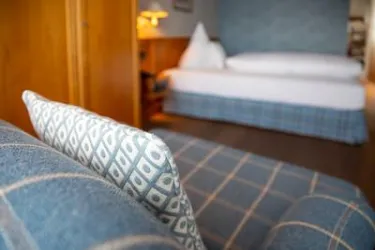 Ein gemütliches Zimmer mit einem Bett im Hintergrund und einer blauen Couch im Vordergrund. Die Einrichtung vermittelt eine einladende Atmosphäre.