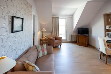 Ein helles Wohnzimmer mit einem Sofa, zwei Sessel und einem Schreibtisch. Große Fenster sorgen für viel Tageslicht und eine gemütliche Atmosphäre.