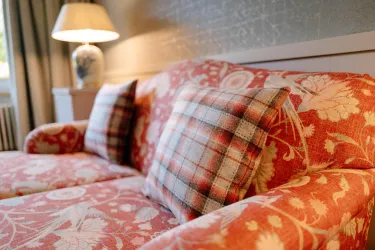 Ein gemütliches Sofa mit floralem Muster und karierten Kissen. Im Hintergrund steht eine Lampe auf einem Beistelltisch.