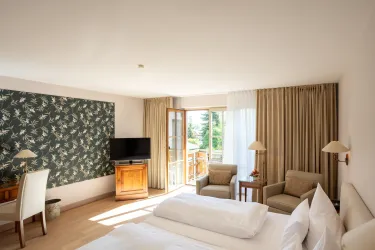 Ein gemütliches Hotelzimmer mit einem großen Doppelbett und einem Fernseher. Große Fenster bieten einen Blick auf den Balkon und den Außenbereich.