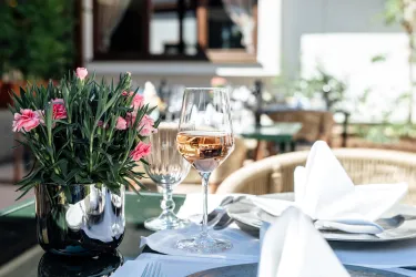 Ein eleganter Tisch in einem Restaurant, gedeckt mit einem Glas Roséwein und einer Blumenvase. Im Hintergrund sind weitere Tische und eine grüne Umgebung zu sehen.