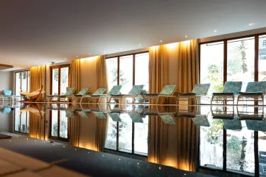 Ein modernes Spa mit Liegen am Beckenrand. Große Fenster lassen viel Licht herein und reflektieren die entspannende Atmosphäre.