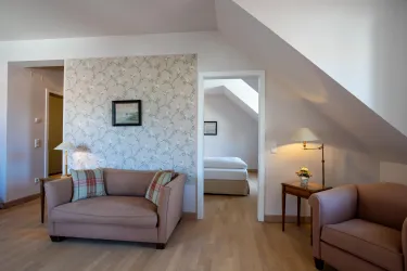 Ein helles Zimmer mit moderner Einrichtung und einem dekorativen Blumenmuster an der Wand. Auf der rechten Seite sind zwei bequeme Sessel und ein Tisch zu sehen.