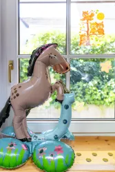 Ein aufgeblasenes Pferd aus Kunststoff steht auf bunten schwimmenden Luftmatratzen. Im Hintergrund sind Fenster mit Blick ins Freie und dekorative Aufkleber zu sehen.