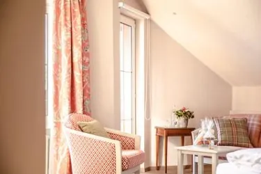 Ein heller Raum mit einem gemütlichen Sessel und einem kleinen Tisch. Die Fenster sind mit bunten Vorhängen dekoriert und es gibt eine Blumenvase auf dem Tisch.
