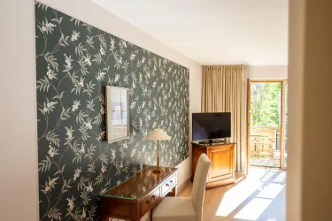 Ein modernes Zimmer mit floralem Design an der Wand. Zu sehen sind ein Schreibtisch, ein Fernseher und ein Balkon mit Tageslicht.