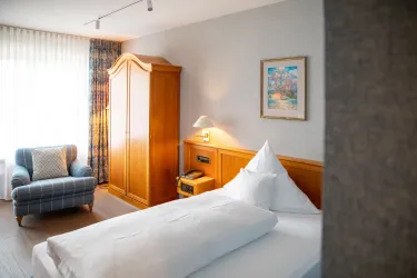Ein gemütliches Schlafzimmer mit einem Einzelbett, einem bequemen Sessel und einem Kleiderschrank aus Holz. Natürliches Licht strömt durch die Fenster und sorgt für eine einladende Atmosphäre.