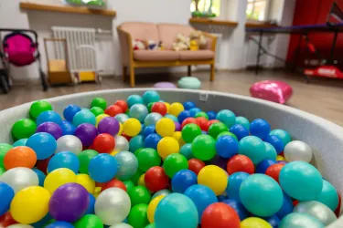 Eine große Schüssel mit bunten Plastikbällen in verschiedenen Farben. Im Hintergrund sind eine Couch und Spielsachen sichtbar.