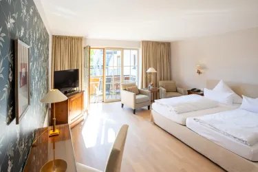 Ein helles Hotelzimmer mit zwei Betten und modernen Möbeln. Große Fenster bieten Zugang zu einem Balkon und viel Tageslicht.
