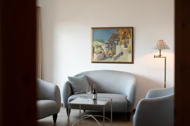 Ein gemütliches Wohnzimmer mit blauen Sofas und einem Tisch. An der Wand hängt ein farbenfrohes Gemälde, und auf dem Tisch stehen eine Flasche Wein und ein Glas.
