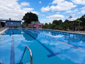 Ein klarer Swimmingpool mit einem blauen Himmel und einigen Wolken. Auf der Seite sind Liegen und Pflanzen zu sehen.