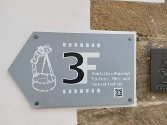 Ein Schild zeigt den Weg zum Deutschen Museum für Foto-, Film- und Fernsehtechnik. Es befindet sich an einer Wand und zeigt eine Pfeilrichtung sowie eine grafische Darstellung.