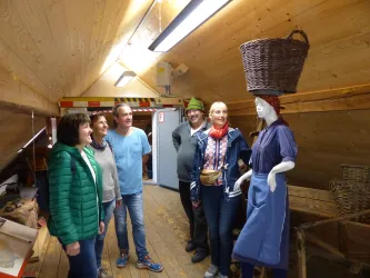 Eine Gruppe von fünf Personen steht in einem Raum in der Samenklenge. In der Mitte befindet sich eine Schaufensterpuppe, die traditionell gekleidet ist und einen Korb auf dem Kopf trägt.