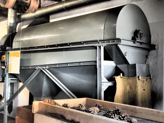 Eine graue Maschine in einem Raum, die für industrielle Zwecke genutzt wird. Sie hat eine spezielle Form und ist mit einem Sack für Materialien ausgestattet. Die Maschine befindet sich in der Alten Samenklenge.