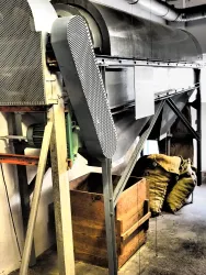 Eine industrielle Maschine steht in einem Raum in der Alten Samenklenge. Im Hintergrund sind Holzsäcke und eine Holzbox sichtbar.