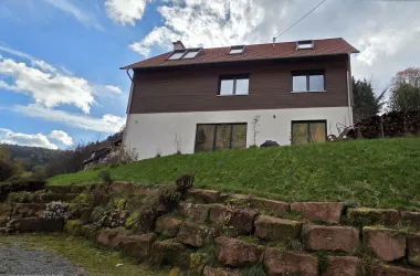 Ein modernes Haus mit brauner Holzverkleidung und großen Fenstern steht auf einem grünen Hügel. Der Himmel ist teils bewölkt mit blauen und grauen Abschnitten.